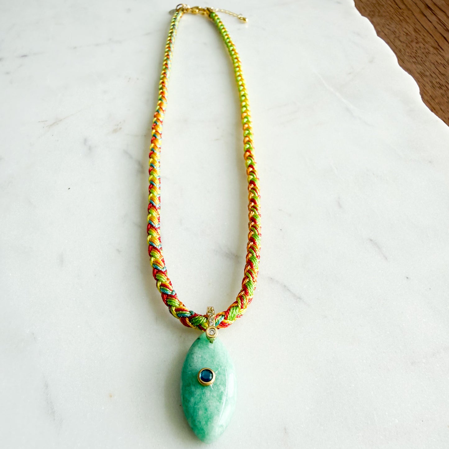 A handmade bohemian-style necklace featuring a vibrant multicolor braided cord and a polished green amazonite pendant with a small blue stone accent, displayed on a white marble surface. The Banks Semi-Precious Stone Braided Rope Necklace - BelleStyle