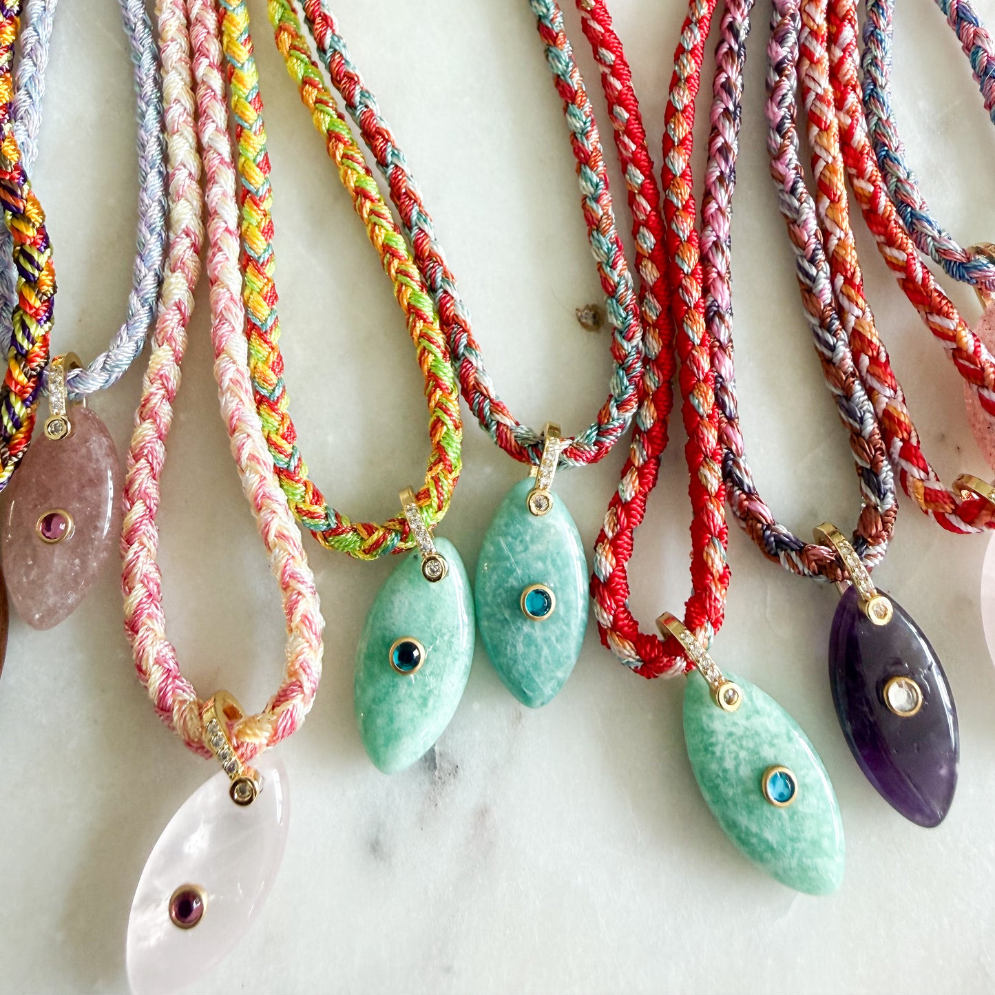 A close-up of several braided cord necklaces with teardrop-shaped stone pendants arranged on a light marble surface. The cords are multicolored, including pink, green, red, blue, and gray, and the pendants are translucent pink, mint green, and purple with small gold eye-like inlays.
The Banks Semi-Precious Stone Braided Rope Necklace- BelleStyle