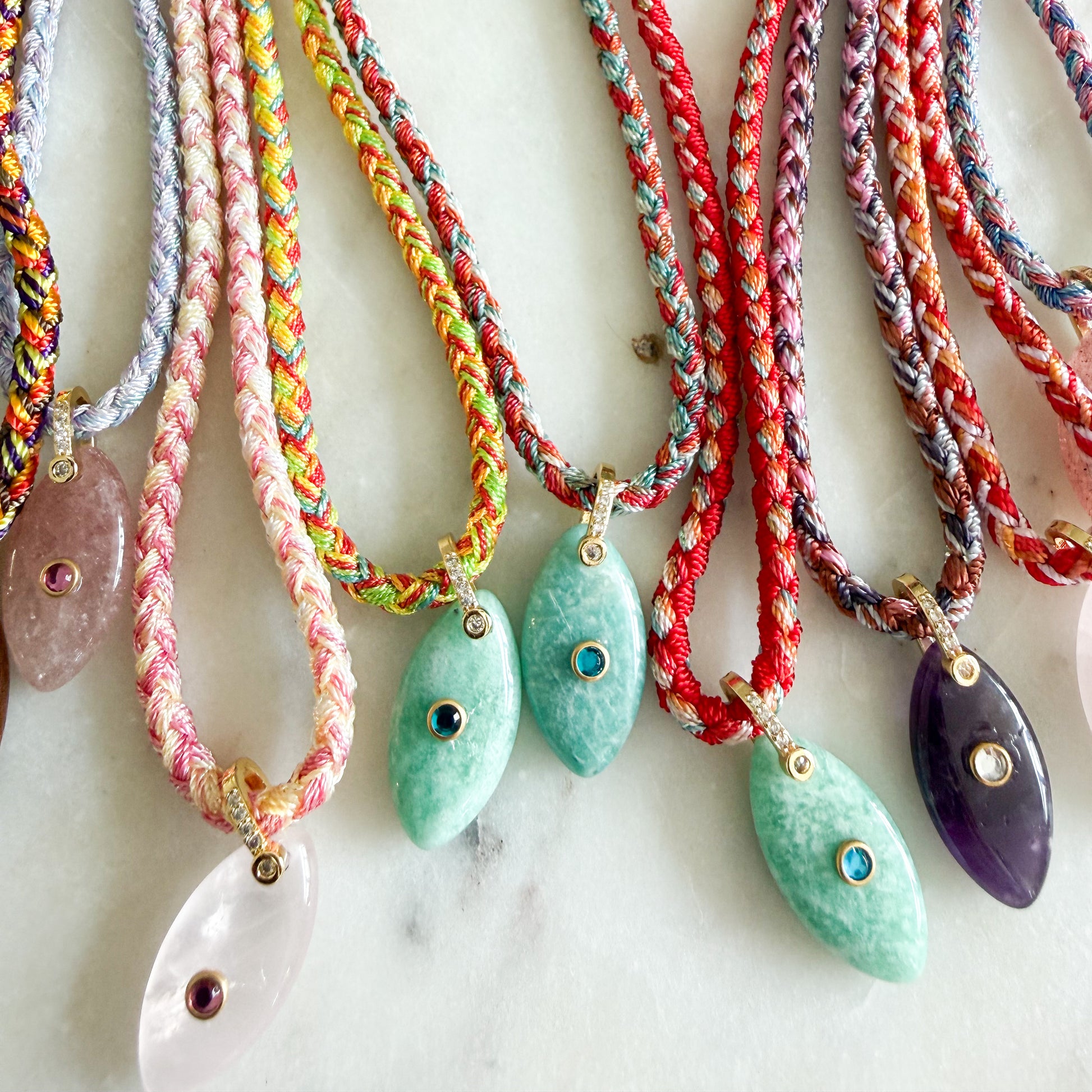 A close-up of several braided cord necklaces with teardrop-shaped stone pendants arranged on a light marble surface. The cords are multicolored, including pink, green, red, blue, and gray, and the pendants are translucent pink, mint green, and purple with small gold eye-like inlays.
The Banks Semi-Precious Stone Braided Rope Necklace- BelleStyle